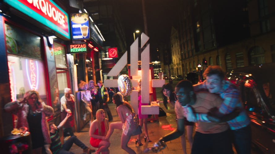Still image from the new Channel 4 idents showing a crowd of people on a night out. One person wearing a short dress rests their foot on another person's lap, while two other people are doing a piggyback on the pavement
