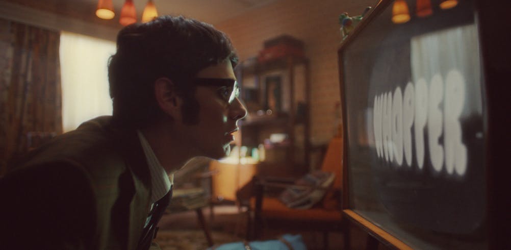 Still image from Burger King's Chicken Royale advert, showing a person with cropped hear, glasses and a retro outfit staring at a 60s TV screen showing the word 'Whopper'