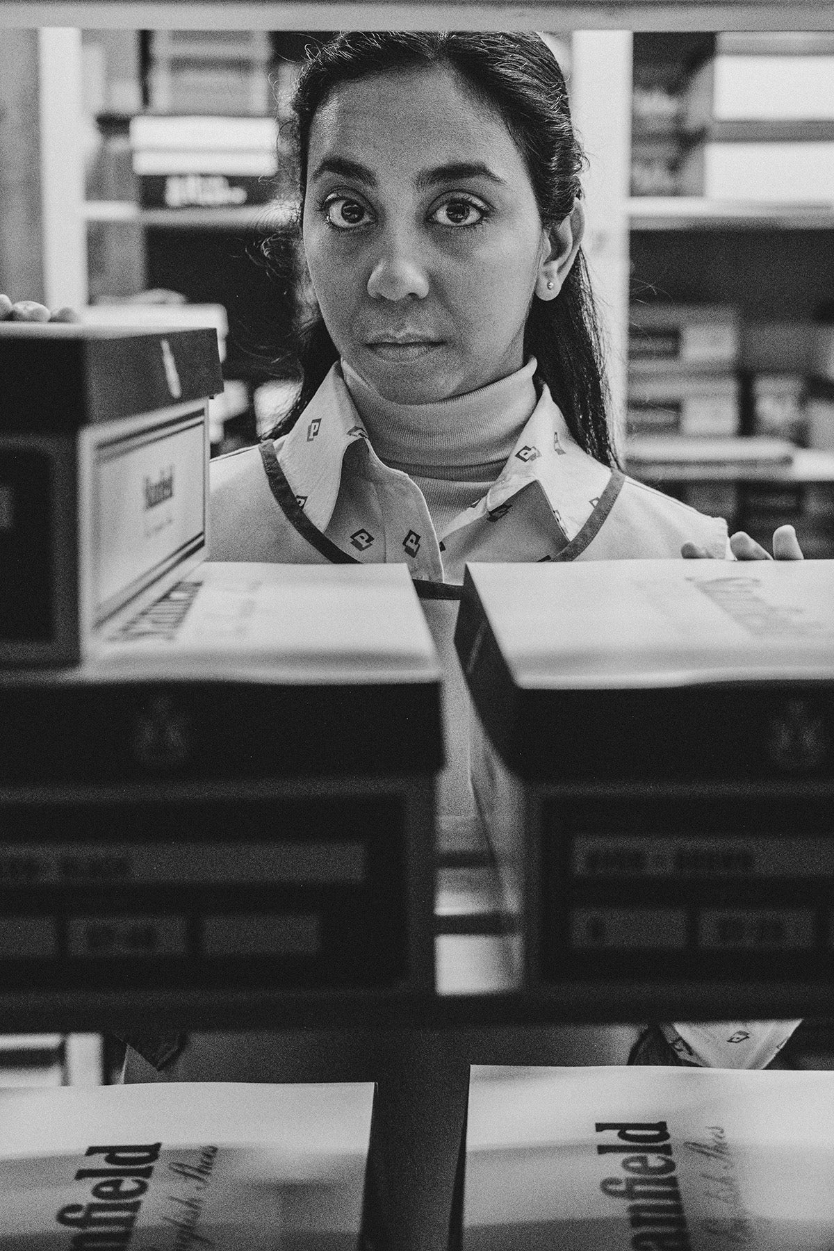 Black and white photograph by David Hurn showing Black Mirror star Anjana Vasan wearing a bib, a collared shirt, and her long hair pinned back, looking at the camera over the top of boxes of files
