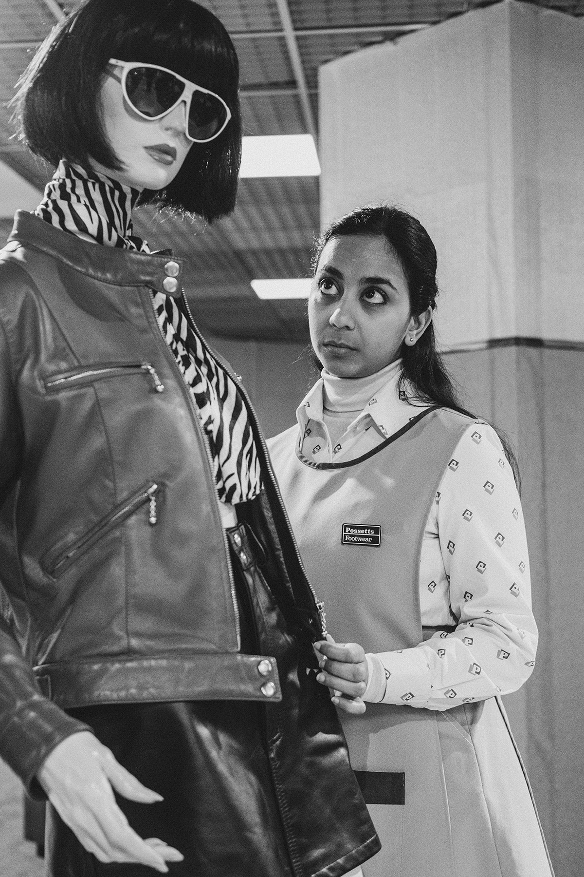 Black and white photograph by David Hurn showing Black Mirror star Anjana Vasan wearing a bib, a collared shirt, and her long hair pinned back, looking up at a mannequin wearing a leather cropped jacket and zebra print scarf