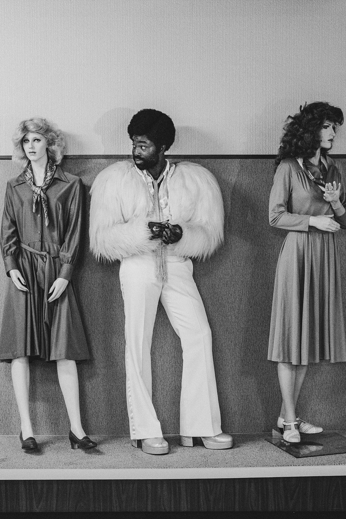 Black and white photograph by David Hurn showing Black Mirror stars Paapa Essiedu in a light feather jacket, slim pale trousers, and a skinny piece of fabric tied around his neck, standin between two mannequins wearing knee-length satin collared dresses