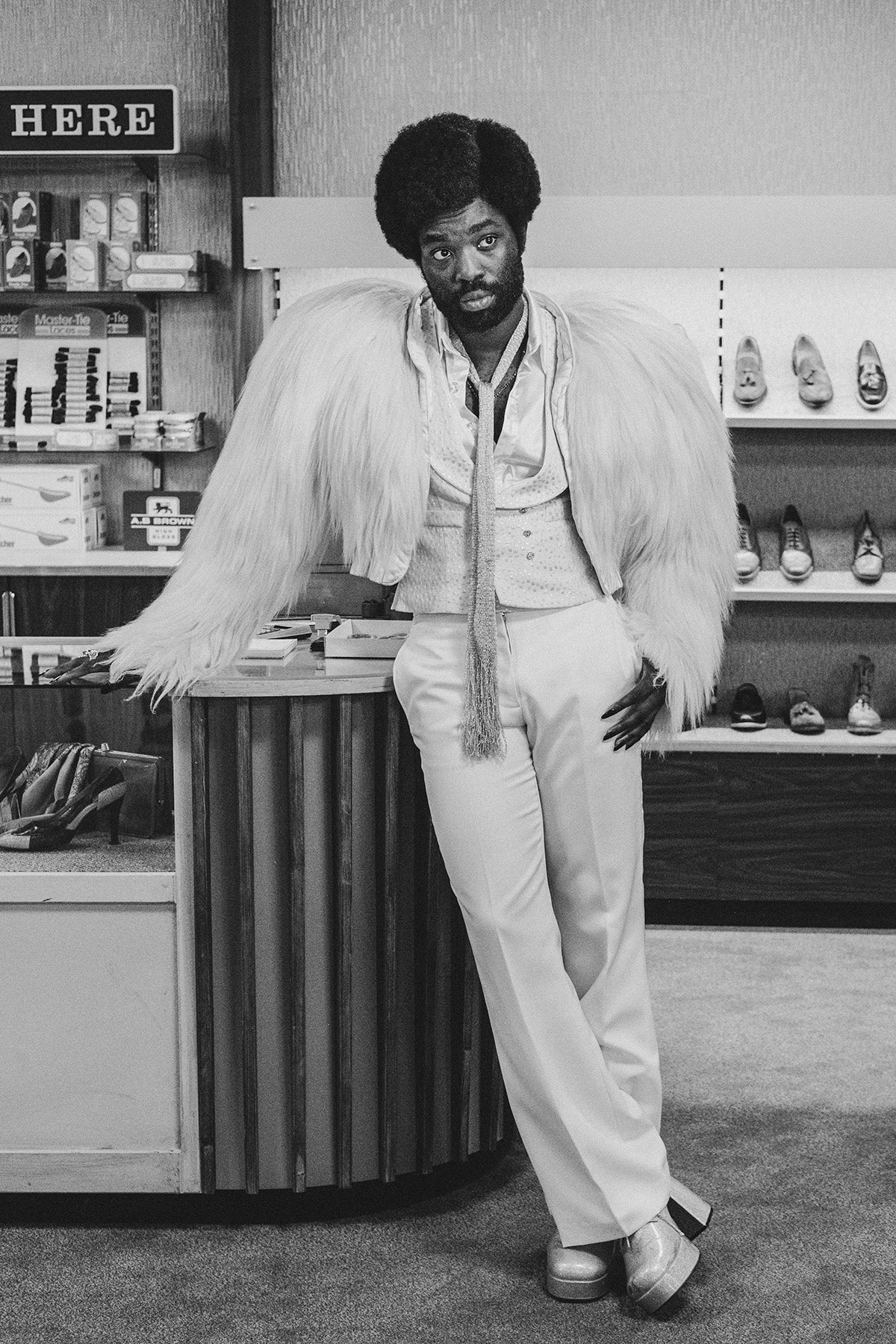 Black and white photograph by David Hurn showing Black Mirror stars Paapa Essiedu in a light feather jacket, slim pale trousers, and a skinny piece of fabric tied around his neck, leaning on a podium in front of racks of shoes