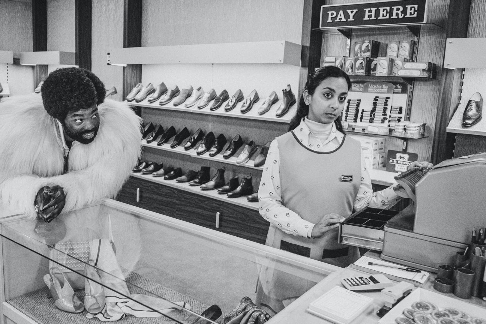 Black and white photograph by David Hurn showing Black Mirror stars Paapa Essiedu in a light feather jacket while leaning on a display case, next to co-star Anjana Vasan wearing a shop assistant's bib and collored shirt, standing over an open till in front of a rack of shoes