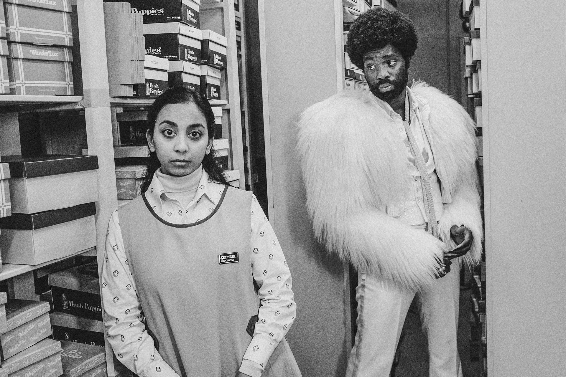 Black and white photograph by David Hurn showing Black Mirror stars Paapa Essiedu in a light feather jacket and slim trousers, looking at co-star Anjana Vasan wearing a shop assistant's bib and collored shirt