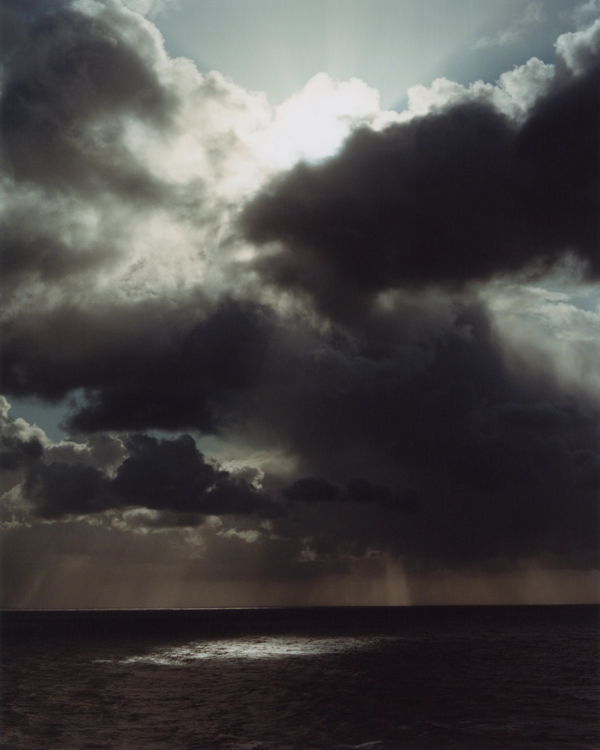 Photograph from Folly by Jamie Murray showing the sea with clouds overhead, with the sun piercing through one, leaving a beam of light on the sea