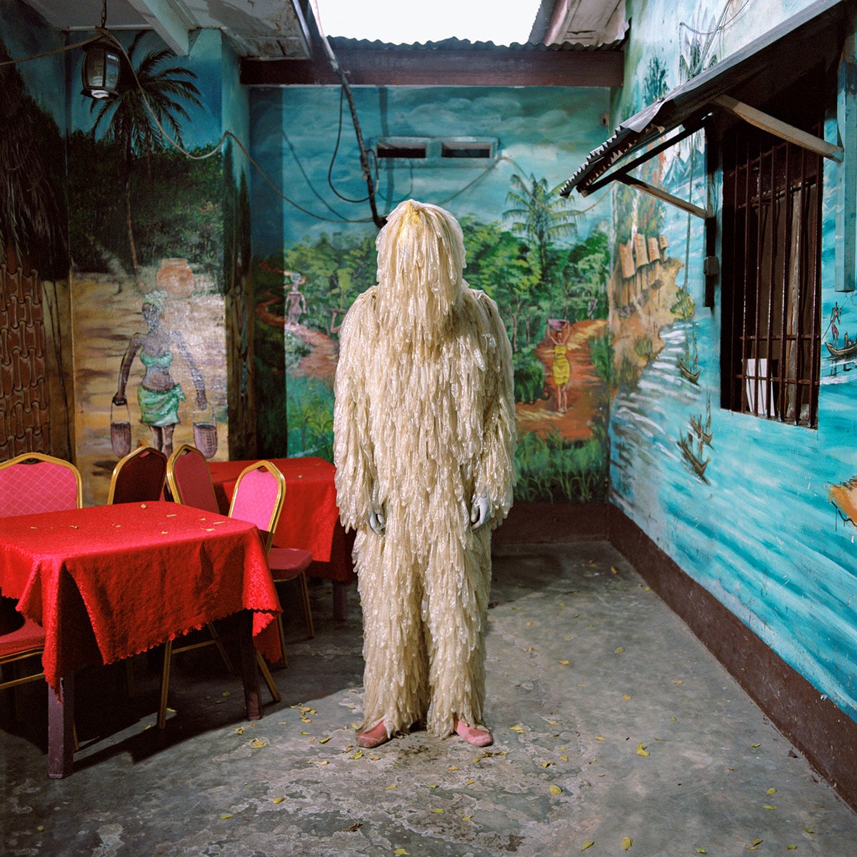 Photo by Colin Delfosse showing a person wearing a beige costume made out of waste, as though covered in layers of long hair, standing in a room with a red table and chairs, and walls painted in a beach scene