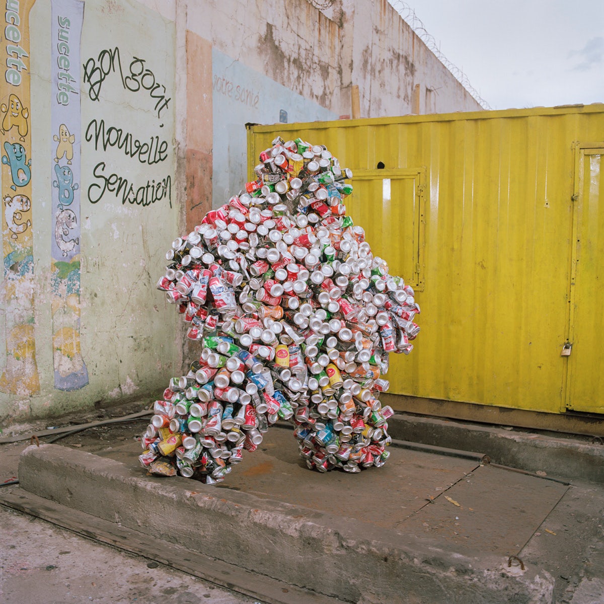 Photo by Colin Delfosse showing a person wearing a bubbly costume made from drinks cans, stood in front of a yellow metal clad container building