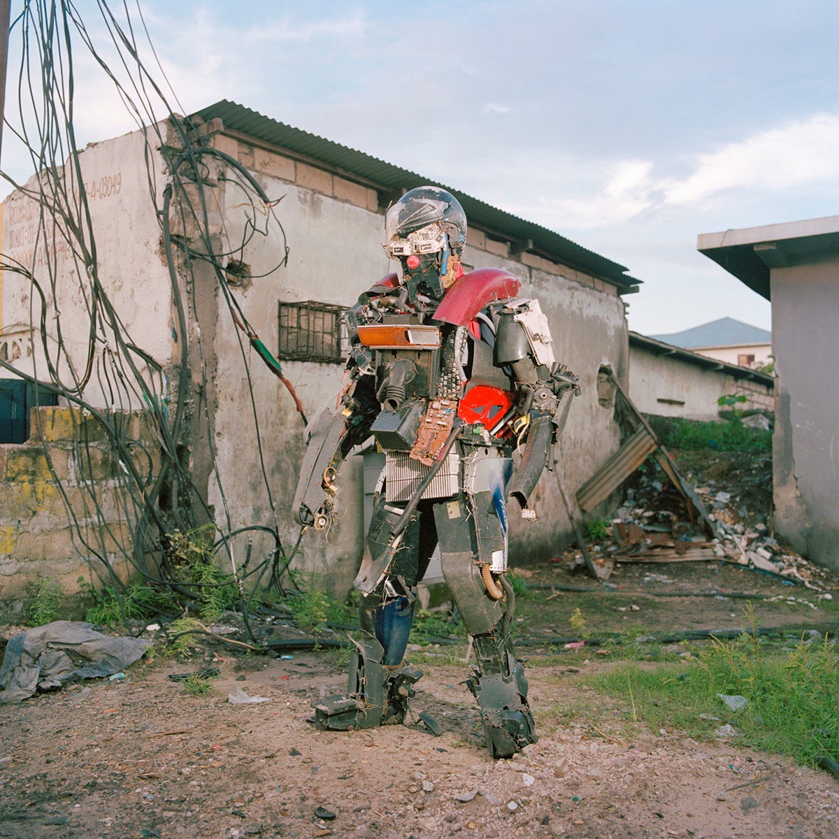 Photo by Colin Delfosse showing a person wearing a costume made out of waste, standing in a yard in front of two low buildings