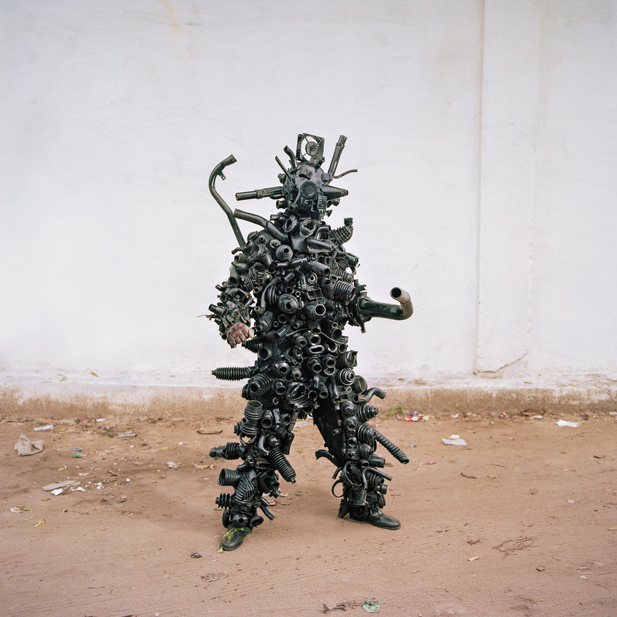 Photo by Colin Delfosse showing a person wearing a dark spiky costume made out of waste such as metallic pipes, stood on rough sandy ground in front of a white wall