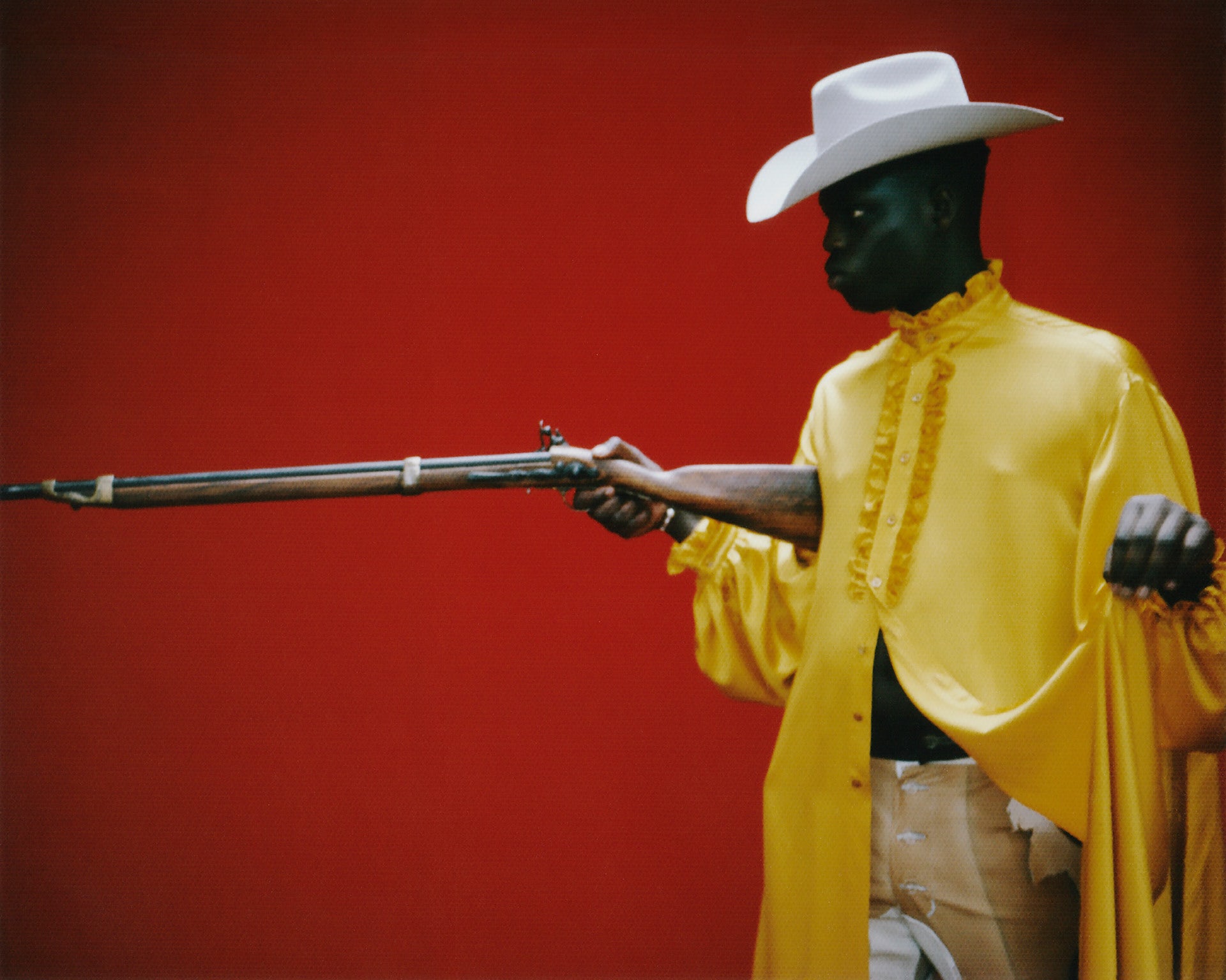 Image by Gabriel Moses of a person wearing a yellow shirt and white cowboy hat pointing a gun to the left of the image against a red backdrop
