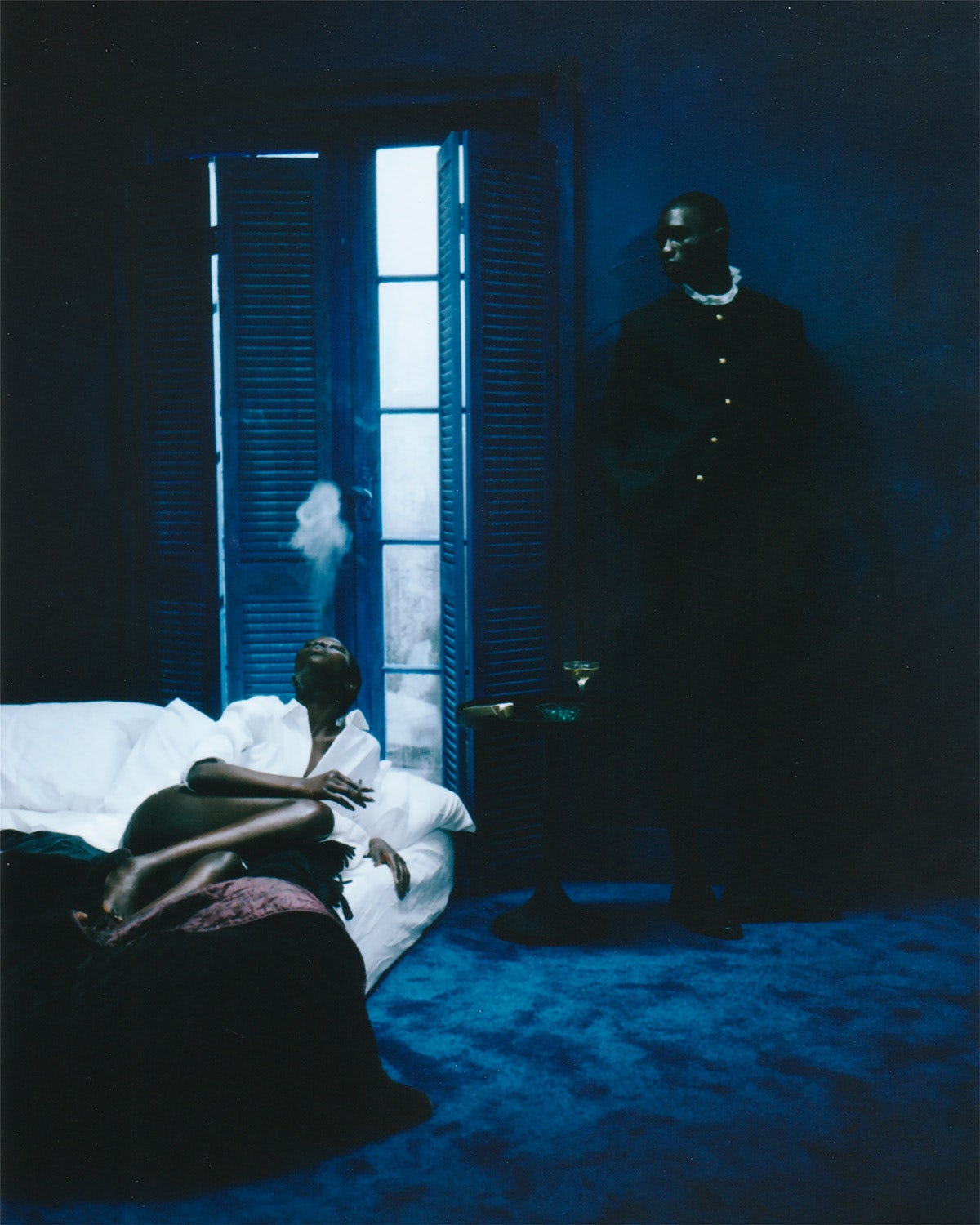 Photograph by Gabriel Moses showing a person reclining on a bed blowing smoke above their head and another person leaning against a wall in a darkened blue room