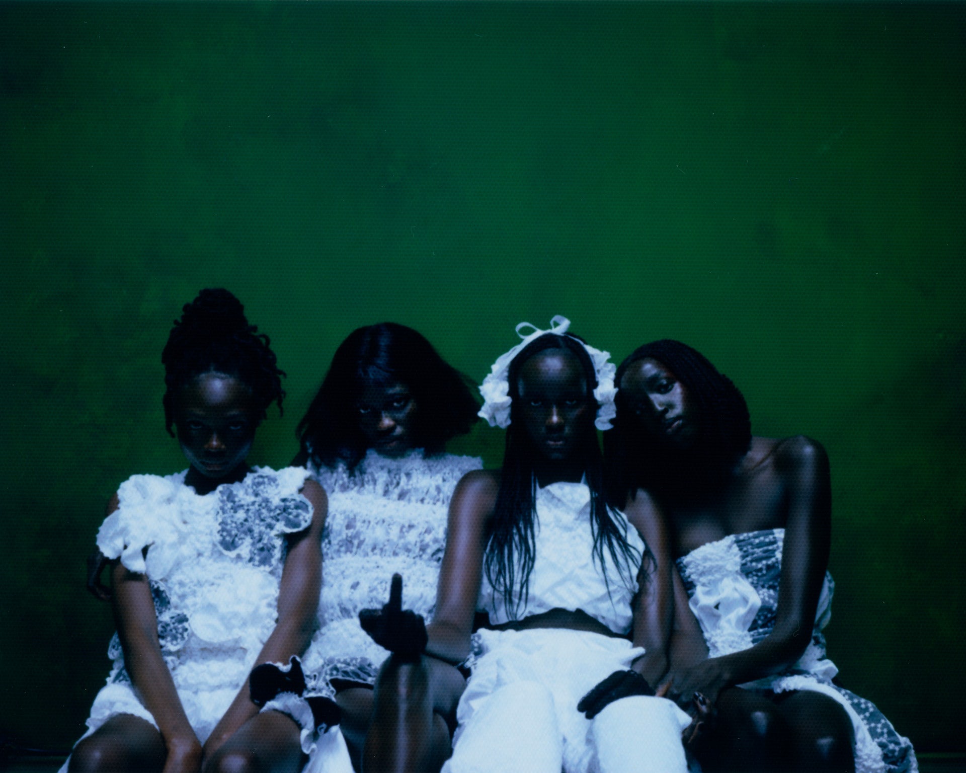 Photograph by Gabriel Moses showing four people leaning against a green backdrop wearing white outfits, with one person holding their middle finger up to the camera